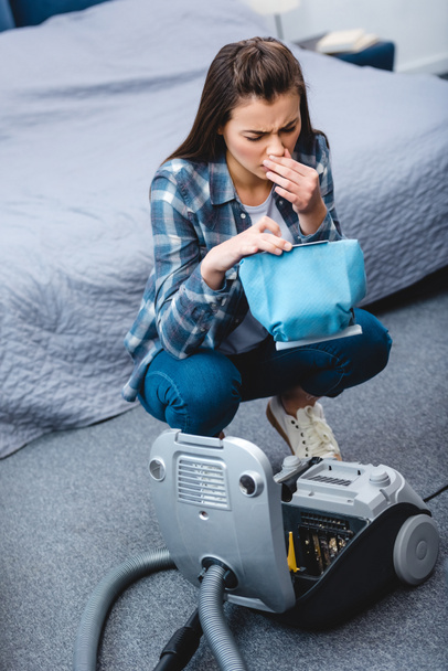 high angle view of young woman with allergy sneezing while holding container from vacuum cleaner with dust   - Photo, Image