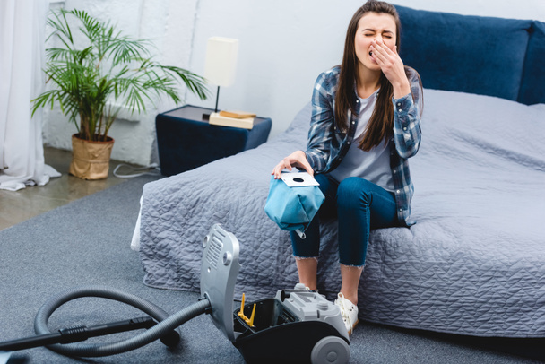 high angle view of woman with allergy holding container from vacuum cleaner with dust and sneezing in bedroom    - Fotó, kép