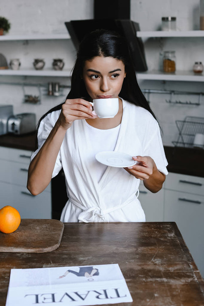 beautiful mixed race girl in white robe drinking coffee in morning in kitchen - Photo, Image