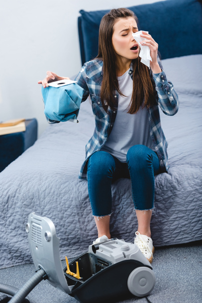 meisje met allergie holding container uit stofzuiger met stof en niezen in slaapkamer  - Foto, afbeelding