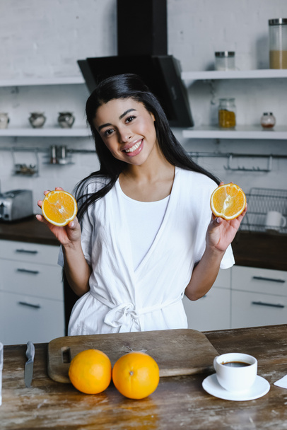 smiling beautiful mixed race girl in white robe holding cut orange and looking at camera in morning in kitchen - Photo, Image