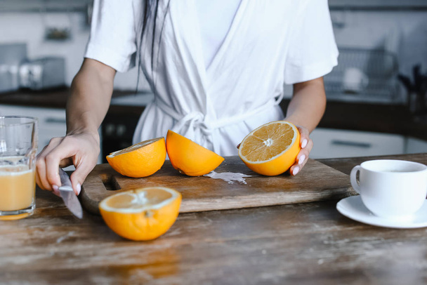 imagen recortada de chica de raza mixta en bata blanca preparando jugo de naranja por la mañana y el corte de naranjas en la cocina
 - Foto, Imagen