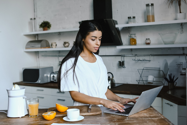 bela menina de raça mista em roupão branco usando laptop de manhã na cozinha
 - Foto, Imagem