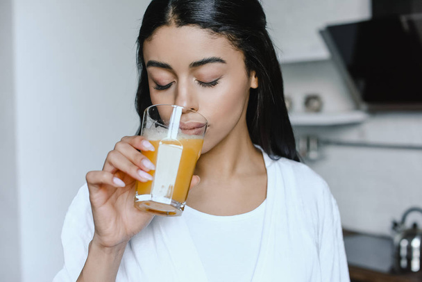 beautiful mixed race girl in white robe drinking fresh orange juice in morning in kitchen with closed eyes - Foto, Bild