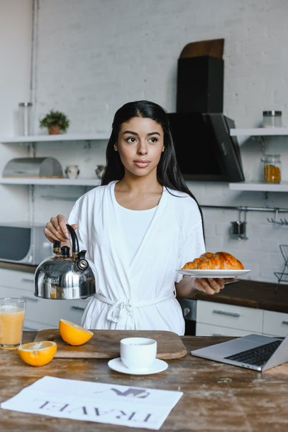 beautiful mixed race girl in white robe holding kettle and plate with croissants in morning in kitchen - Zdjęcie, obraz