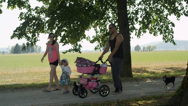 Χαμογελώντας μπαμπάς τατουάζ σπρώχνοντας νεογέννητο μωρό τους σε ένα ροζ καροτσάκι μέσα από το υπέροχο πάρκο μαζί με την ενεργητική κόρη του, όμορφη γυναίκα και χαριτωμένο μικρό σκυλί. Αξιολάτρευτη σύγχρονη οικογένεια απολαμβάνει την άνοιξη. - Φωτογραφία, εικόνα