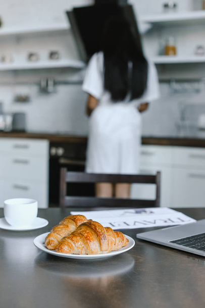 Hátulnézet kevert lány fehér palástban állva közelében tűzhely reggel konyhában, előtérben a croissant - Fotó, kép
