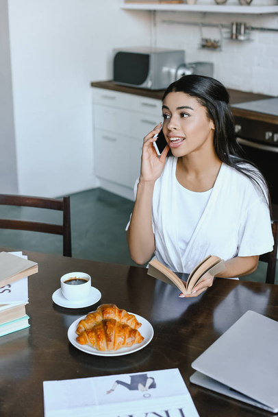 mosolyogva vonzó vegyes verseny lány fehér köntöst konyhában reggel beszél a smartphone és a gazdaság könyv - Fotó, kép