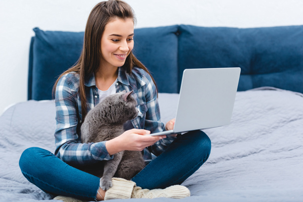 smiling young woman using laptop while sitting on bed with cute british shorthair cat  - Photo, Image