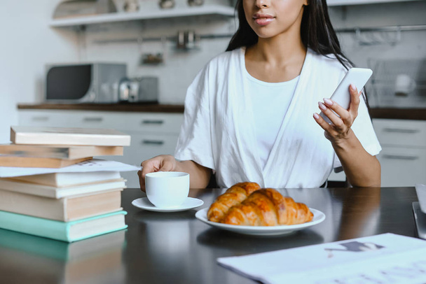 levágott kép lány kevert állomány smartphone és csésze kávé reggel konyha fehér köntöst - Fotó, kép