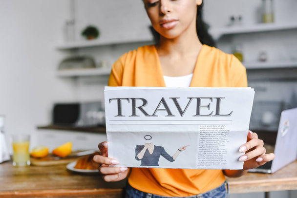selective focus of beautiful mixed race girl in orange shirt holding travel newspaper in kitchen - Photo, Image