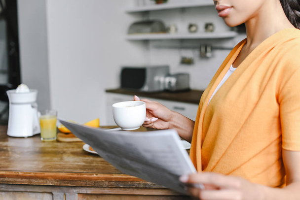 abgeschnittenes Bild eines gemischten Rassenmädchens in orangefarbenem Hemd, das eine Tasse Kaffee hält und in der Küche Zeitung liest - Foto, Bild