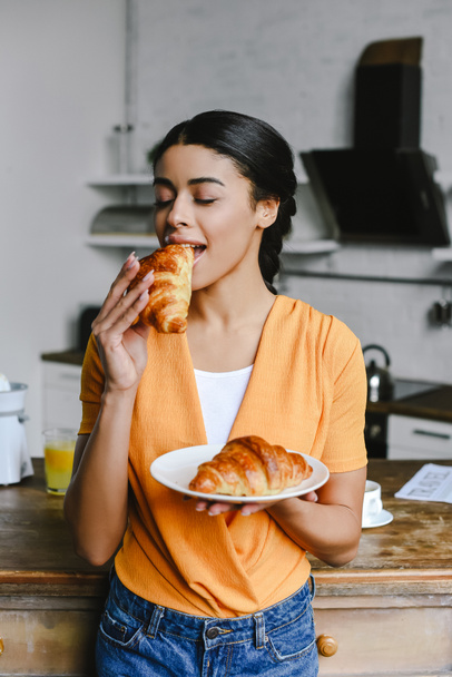 beautiful mixed race girl in orange shirt eating croissant on breakfast in kitchen - Foto, Bild