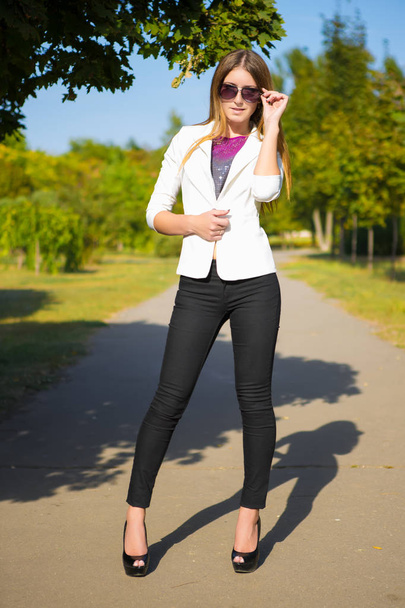 atractiva joven en pantalones negros y chaqueta blanca posando en la carretera en el parque
 - Foto, imagen