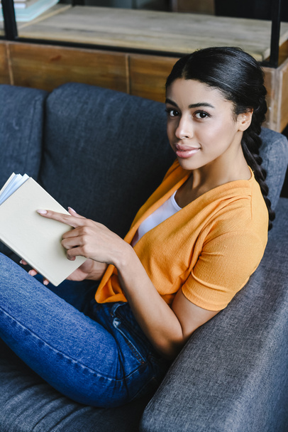 hoge hoekmening van mooi gemengd ras meisje in oranje shirt boek houden en kijken naar camera in woonkamer - Foto, afbeelding