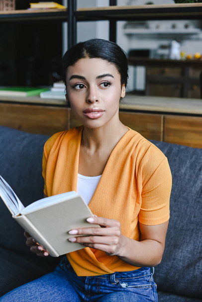hermosa chica de raza mixta en camisa naranja sosteniendo libro en la sala de estar y mirando hacia otro lado
 - Foto, Imagen