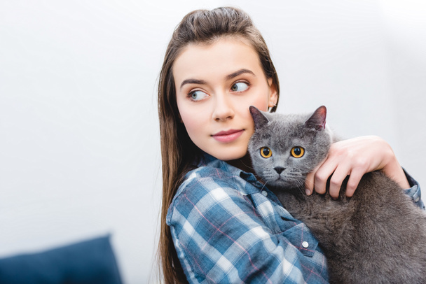girl holding british shorthair cat and looking away at home - Fotó, kép