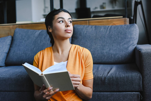 beautiful mixed race girl in orange shirt holding book in living room and looking away - Photo, Image