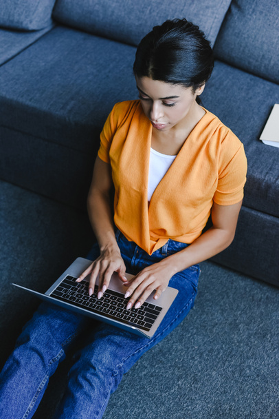 high angle view of beautiful mixed race girl in orange shirt sitting on floor and using laptop in living room - Fotoğraf, Görsel