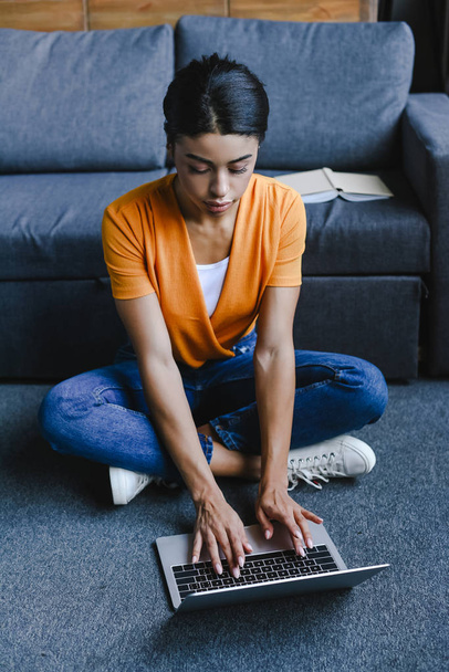hermosa chica de raza mixta en camisa naranja sentado en el suelo y el uso de ordenador portátil en la sala de estar
 - Foto, Imagen