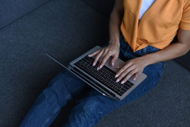обрезанное изображение девушки смешанной расы в оранжевой рубашке с помощью ноутбука в гостиной
 - Фото, изображение