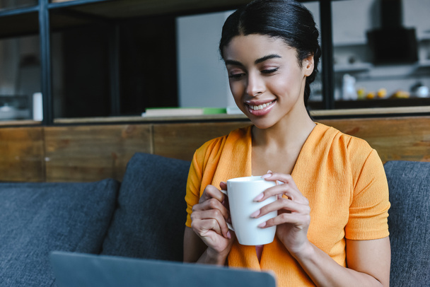 smiling beautiful mixed race girl in orange shirt using laptop and holding cup tea in living room - Foto, Imagem