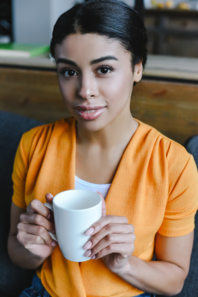 narancssárga inget gazdaság csésze teát, és látszó-on fényképezőgép otthon szép kevert lány portréja - Fotó, kép