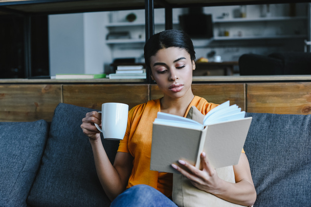 attractive mixed race girl in orange shirt holding cup of tea and reading book on sofa at home - Photo, Image