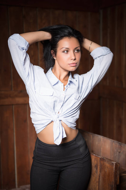 attractive brunette woman in white shirt and black pants in front of wooden wall looking away - Fotografie, Obrázek