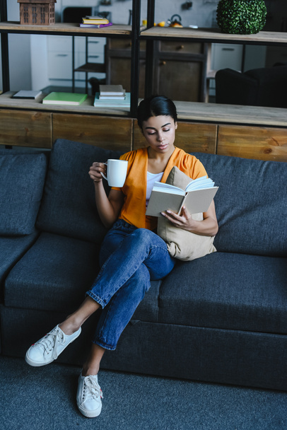 vista de ángulo alto de hermosa chica de raza mixta en camisa naranja sosteniendo taza de té y libro de lectura en el sofá en casa
 - Foto, Imagen