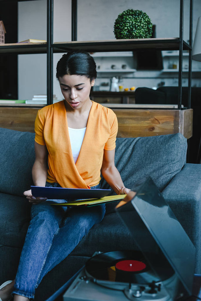 attractive mixed race girl in orange shirt choosing vinyl on sofa in living room - Photo, Image