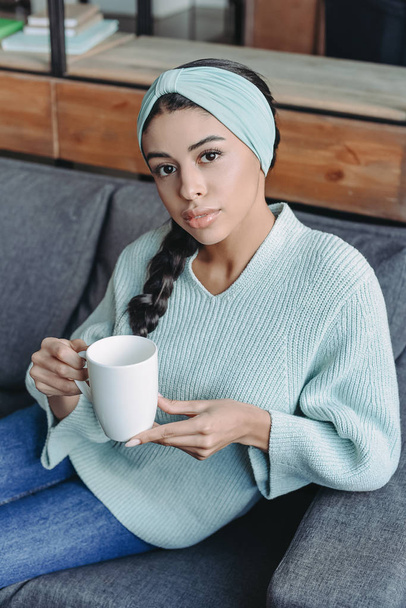 attractive mixed race girl in turquoise sweater and headband sitting on sofa and holding cup of tea in living room - Fotoğraf, Görsel