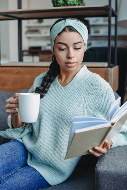 vonzó kevert lány türkizkék pulóver és fejpánt ül a kanapén, és a gazdaság csésze tea és olvasó könyv nappali - Fotó, kép