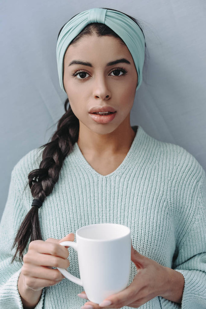 atraktivní smíšený závod dívka v tyrkysový svetr a čelenky držící šálek čaje doma a při pohledu na fotoaparát - Fotografie, Obrázek