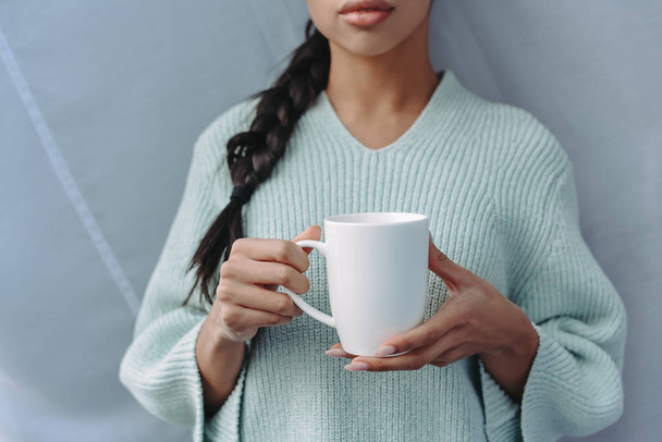 imagen recortada de chica de raza mixta en suéter turquesa sosteniendo taza de té en casa
 - Foto, Imagen