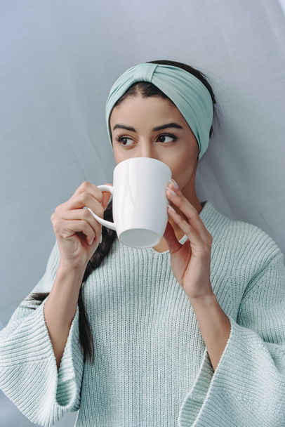 привлекательная девушка смешанной расы в бирюзовом свитере и ободке, пьющая чай дома
 - Фото, изображение