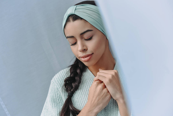 attractive mixed race girl in turquoise sweater and headband standing with closed eyes at home - Photo, Image