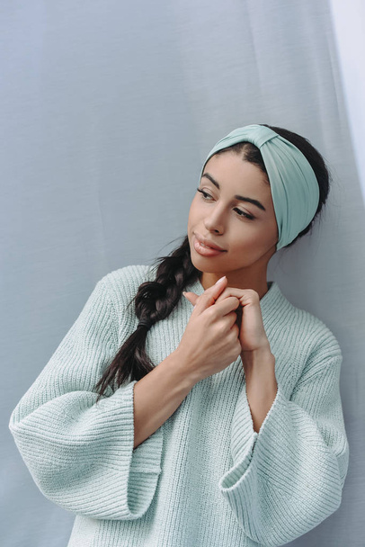attractive mixed race young woman in turquoise sweater and headband at home and looking away  - Zdjęcie, obraz
