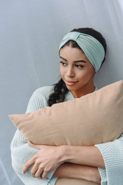 hermosa chica de raza mixta en suéter turquesa y diadema abrazando almohada y mirando hacia otro lado
 - Foto, Imagen