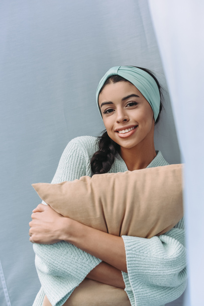 smiling beautiful mixed race girl in turquoise sweater and headband hugging pillow and looking at camera at home - Photo, Image