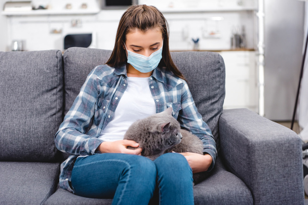 νεαρή γυναίκα στην ιατρική μάσκα κρατώντας γάτα και που πάσχουν από αλλεργία στο σπίτι   - Φωτογραφία, εικόνα