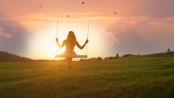 GOLDEN LIGHT SILHOUETTE: Unrecognizable girl in white dress swaying on a tree swing on peaceful evening. Lady sitting on a wooden swing and looking at golden sunset. Young woman swinging at sunrise - Photo, Image