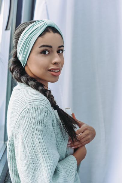 beautiful mixed race girl in turquoise sweater and headband holding book and looking at camera at home - Foto, Imagen
