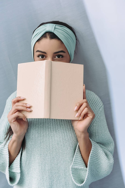 hermosa chica de raza mixta en suéter turquesa y diadema mirando hacia fuera del libro en casa
 - Foto, Imagen