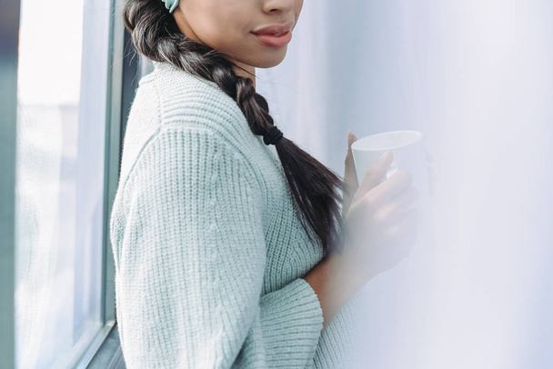 imagen recortada de chica de raza mixta en suéter turquesa sosteniendo taza de té en casa
 - Foto, Imagen