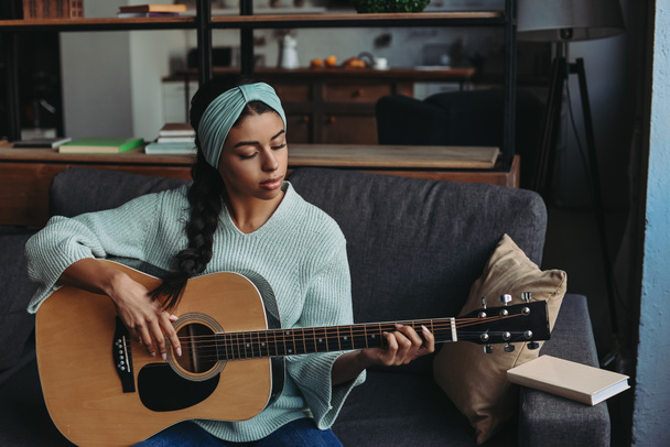 gyönyörű kevert lány türkizkék pulóver és akusztikus gitáron játszik otthon a kanapén fejpánt - Fotó, kép