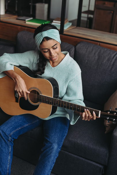 vista de ángulo alto de hermosa chica de raza mixta en suéter turquesa y diadema tocando la guitarra acústica en el sofá en casa
 - Foto, imagen