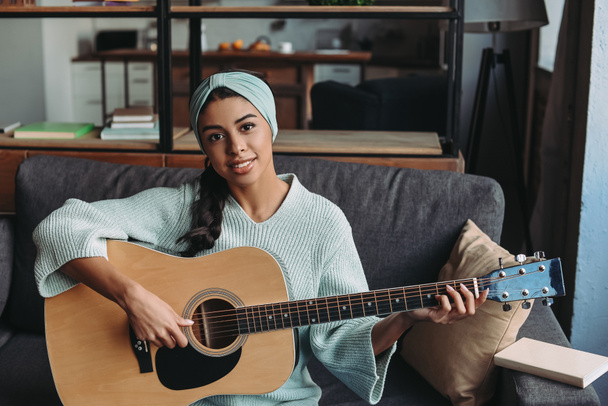 attractive mixed race girl in turquoise sweater and headband playing acoustic guitar on sofa at home and looking at camera - Photo, Image