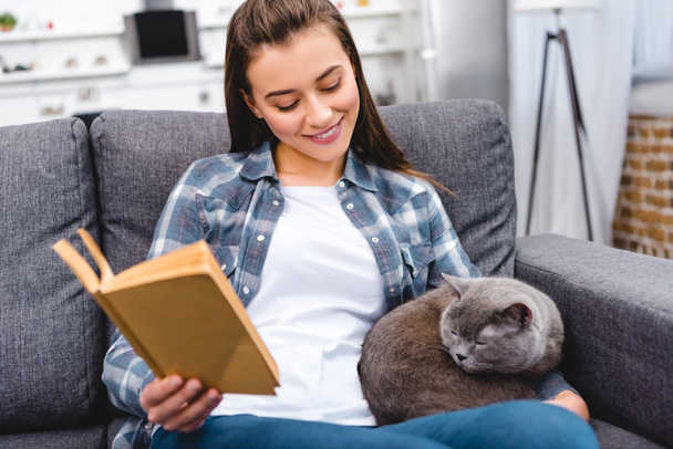 smiling young woman holding book and looking at cute grey cat on sofa - Fotó, kép
