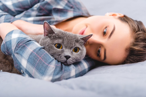 beautiful happy girl lying on bed with british shorthair cat - Fotó, kép